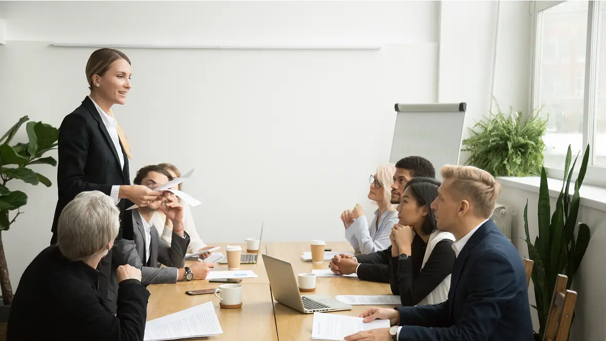 A team meeting around a conference table, led by a woman presenting